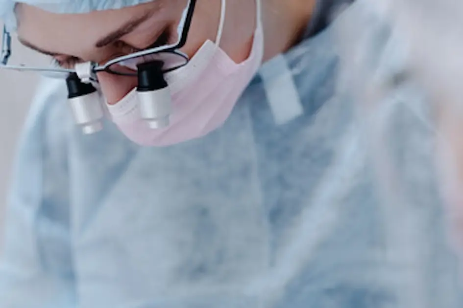 Odontologia dentista O Barco Valdeorras Un cirujano con lupas y una mascarilla quirúrgica se concentra en un procedimiento. El individuo está vestido con una bata quirúrgica y un gorro, lo que indica un ambiente estéril, probablemente dentro de un quirófano.