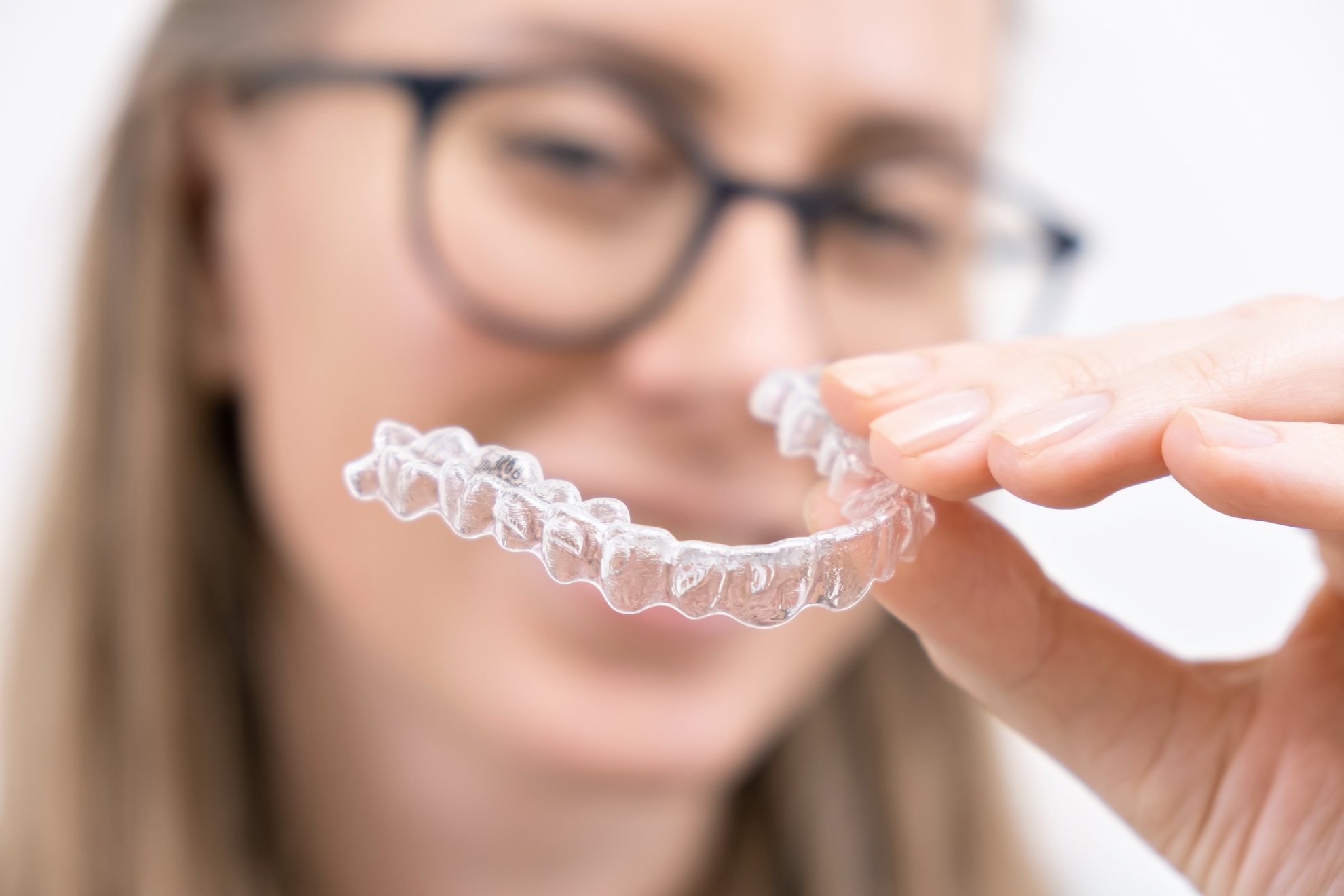 dentista o barco valdeorras Una persona con gafas sostiene un alineador dental transparente enfocado, mostrando su diseño transparente. El alineador se sostiene entre el pulgar y el índice, con la persona visible pero borrosa en el fondo.